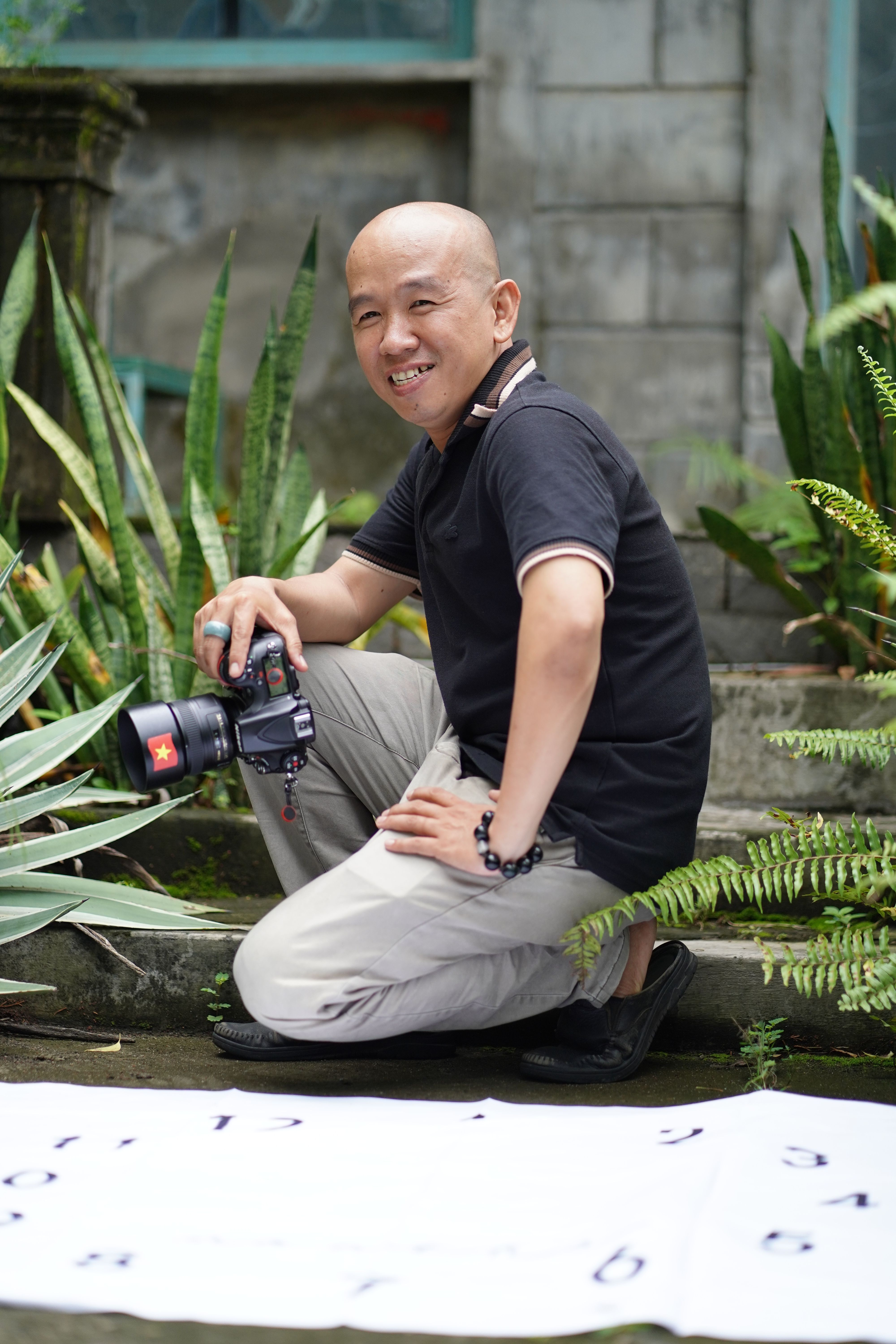 Smiling man holding a camera in an outdoor setting with lush green plants.