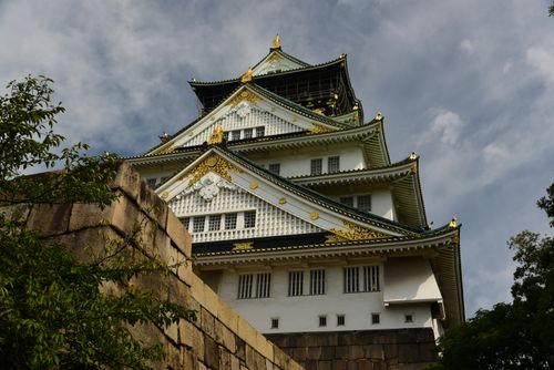 Free A low-angle view of the historic Osaka Castle framed by lush greenery under a cloudy sky. Stock Photo