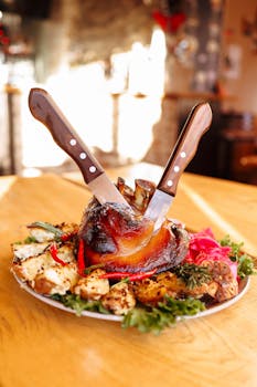 A delicious roasted pork knuckle served with grilled vegetables and herbs on a wooden table in a warm setting.