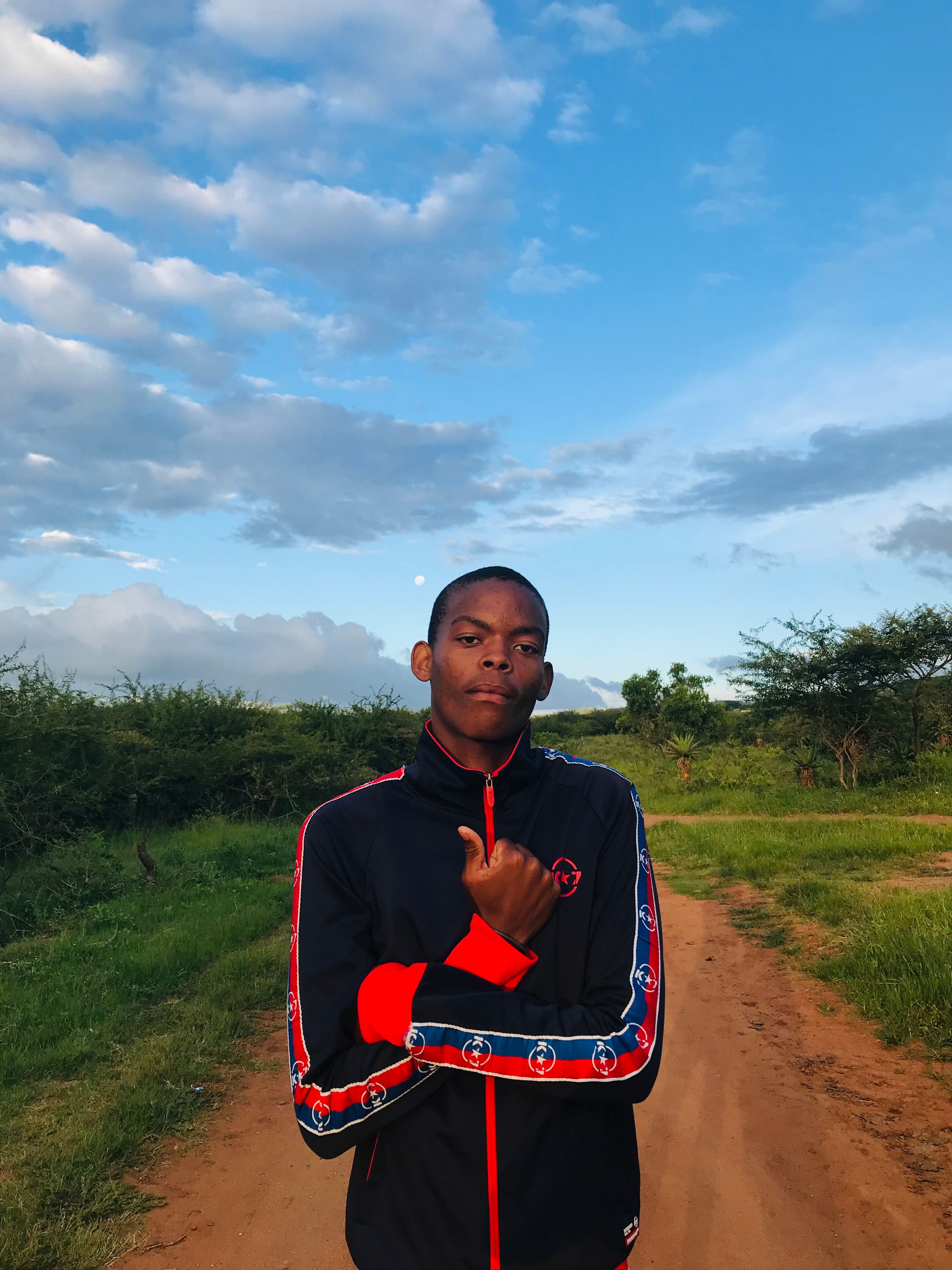 Young man standing on dirt road under bright blue sky, pointing to himself.
