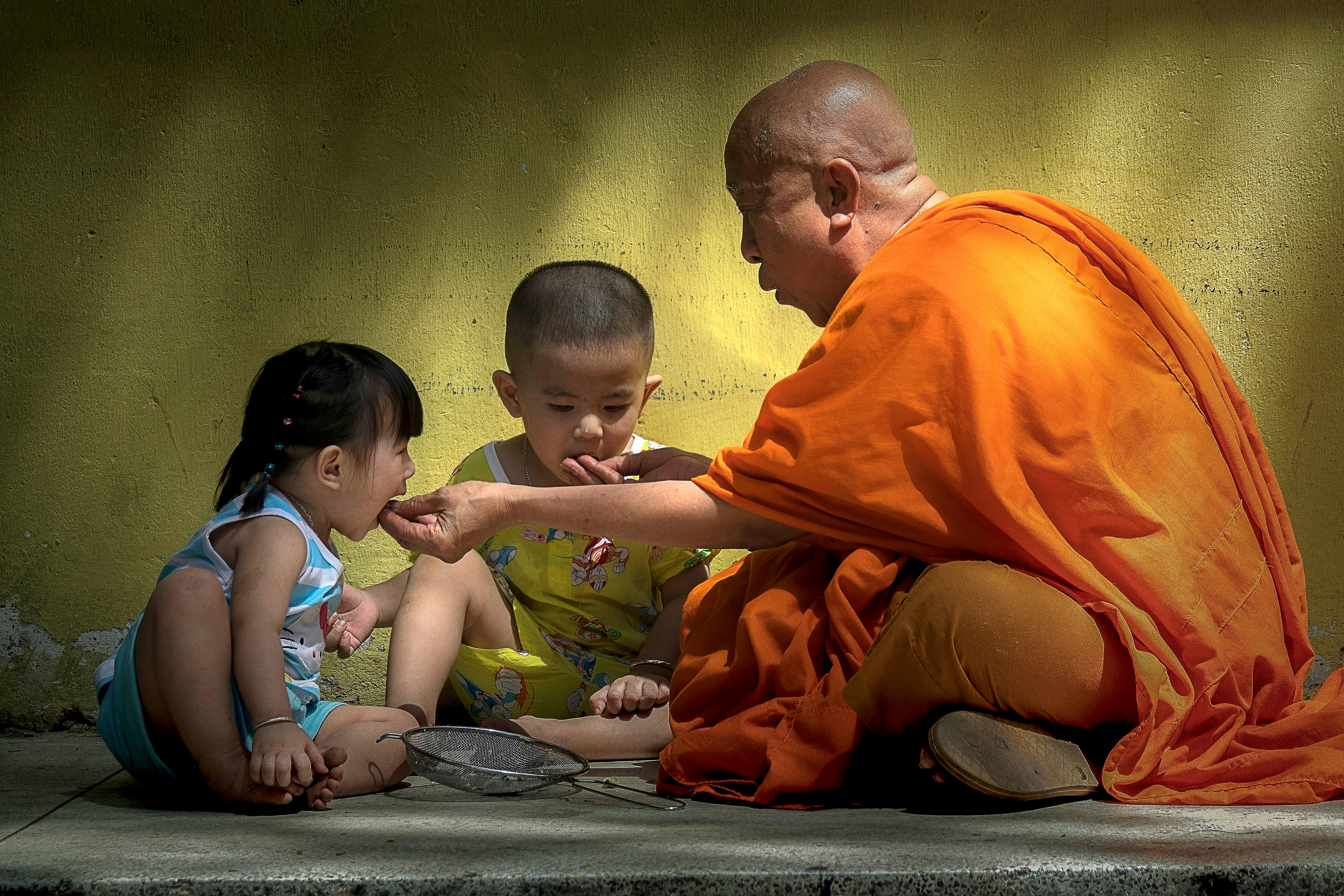 A Buddhist monk shares wisdom with two children seated on the ground in sunlight.