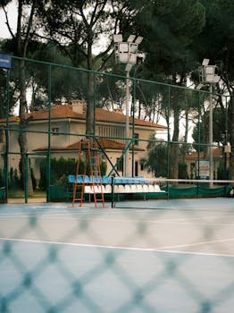 An empty outdoor tennis court surrounded by trees, seating, and floodlights, perfect for sports venues.