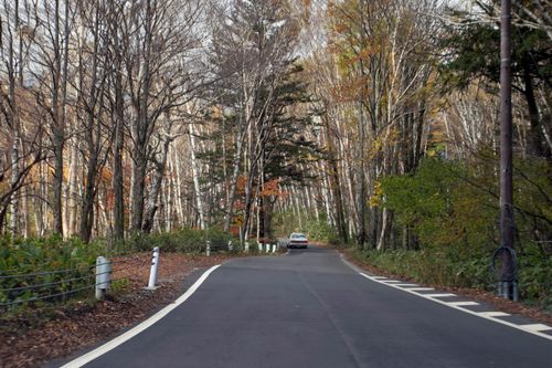 Free Empty road winding through a serene autumn forest with colorful foliage and a distant car. Stock Photo