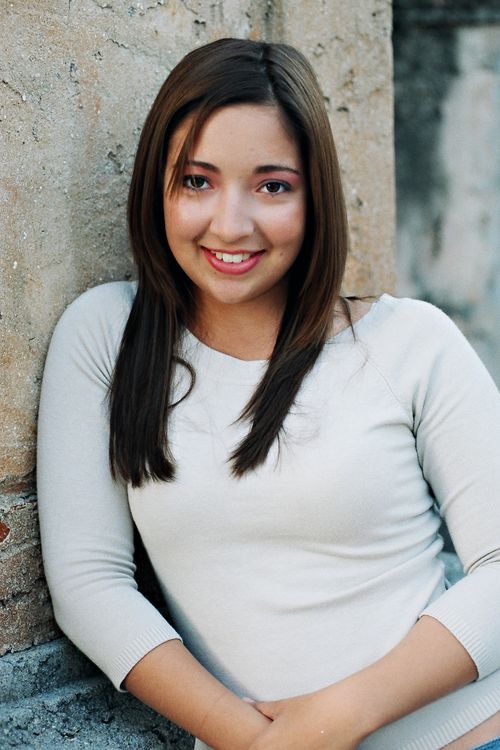 Free Portrait of a young woman with brown hair smiling, leaning against a stone wall outdoors. Stock Photo