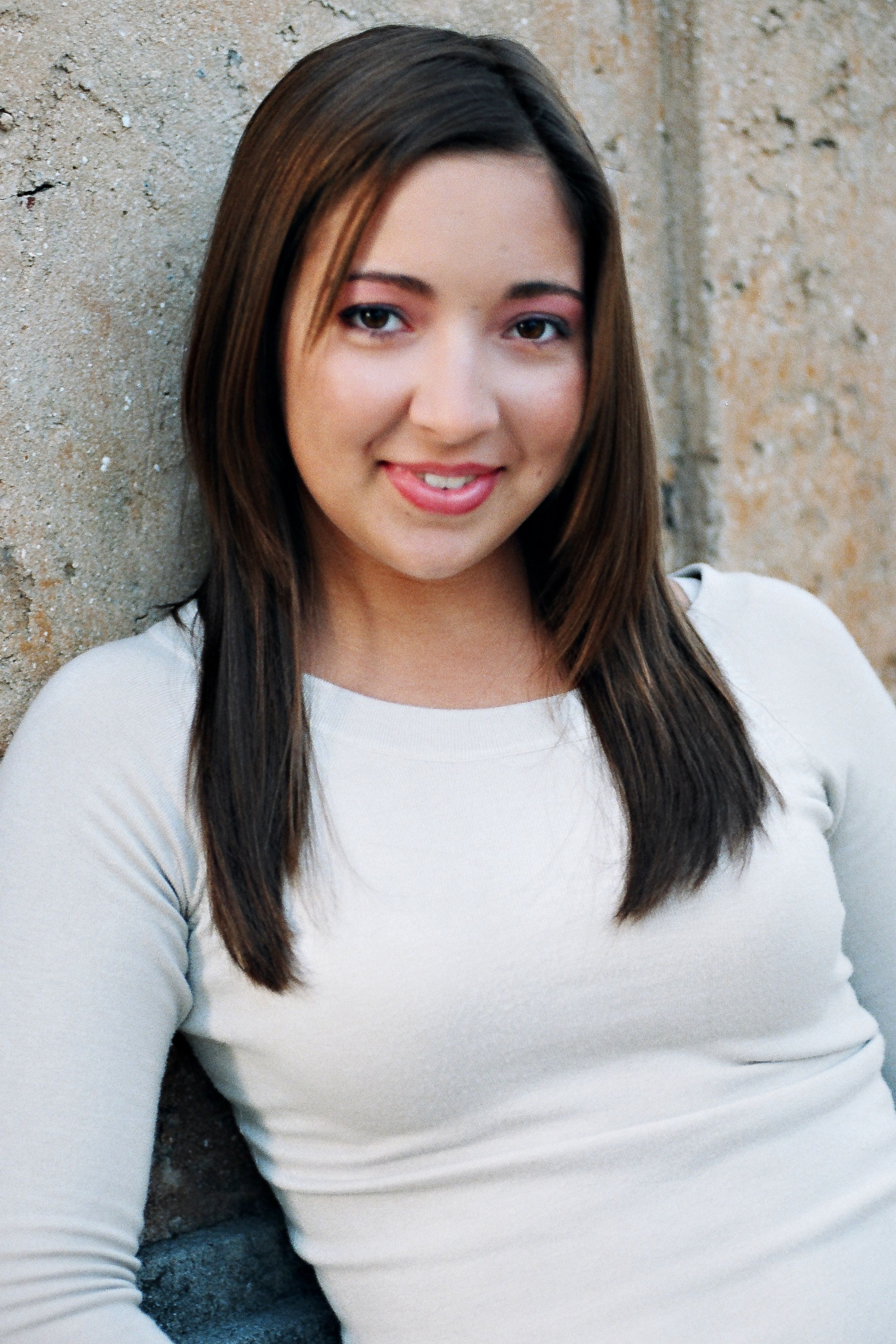 Smiling young woman in white shirt leaning against a textured wall outdoors.