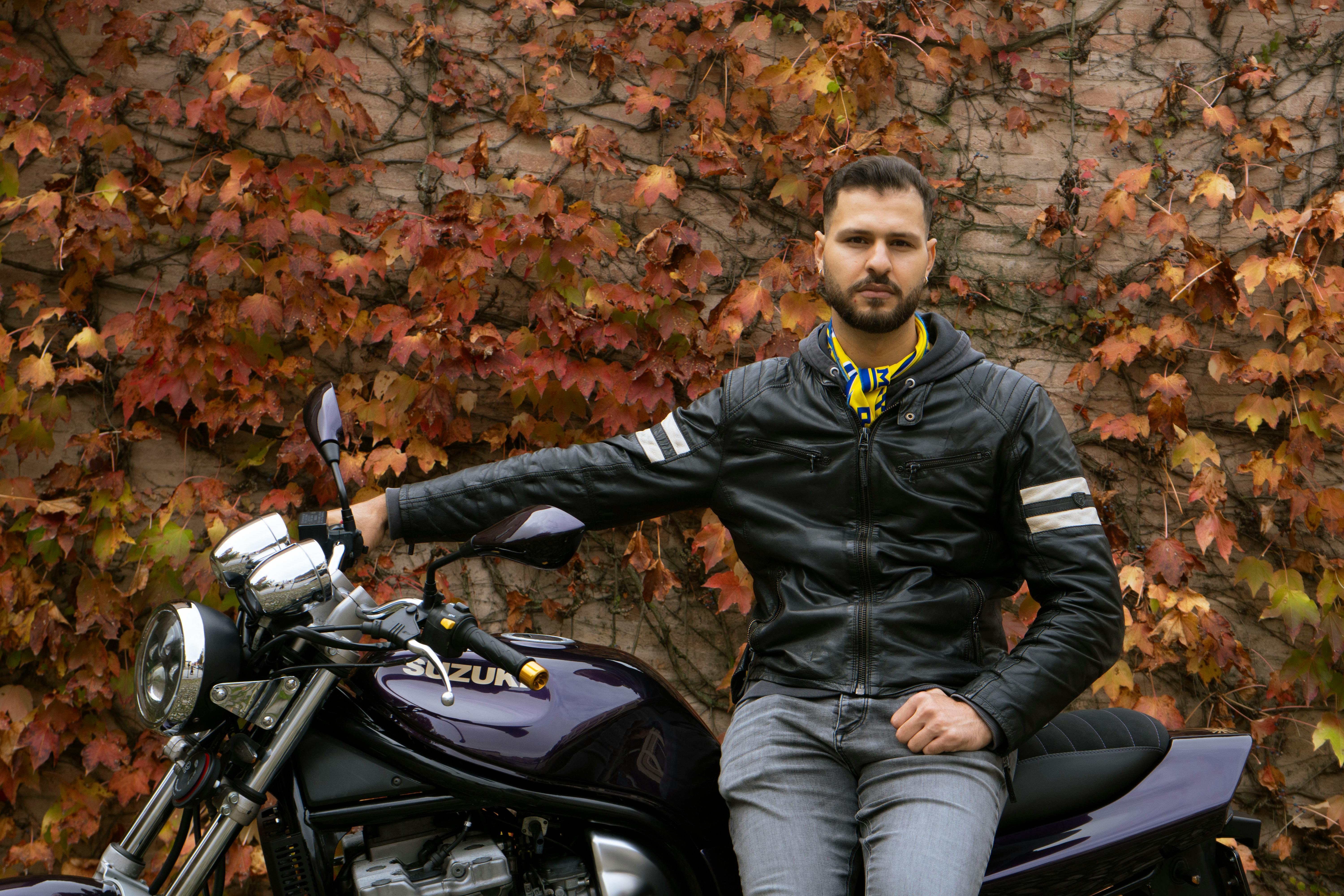 Adult man in leather jacket poses with motorcycle in autumn setting.