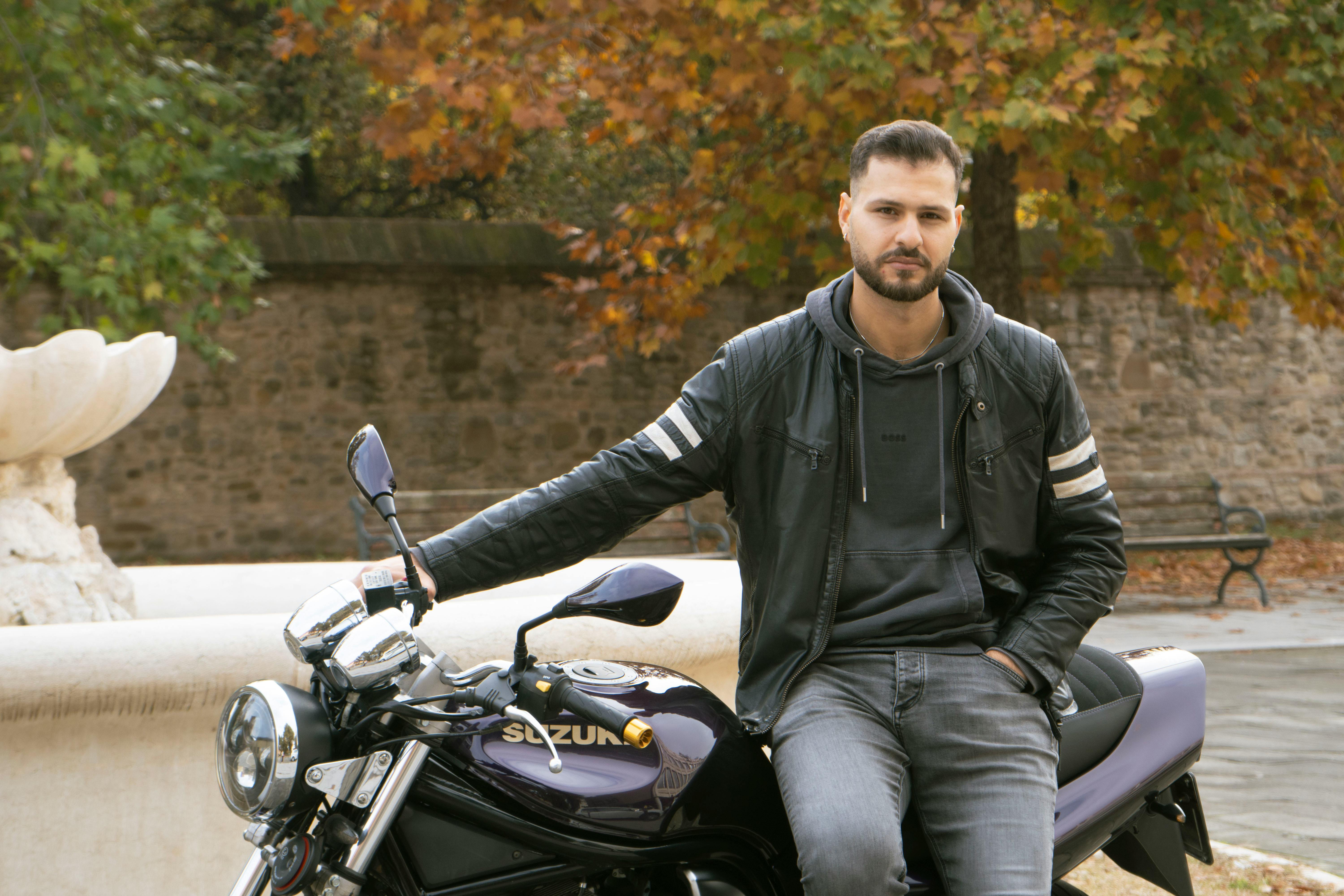 A man in a leather jacket posing with a Suzuki motorcycle in an autumn park setting.