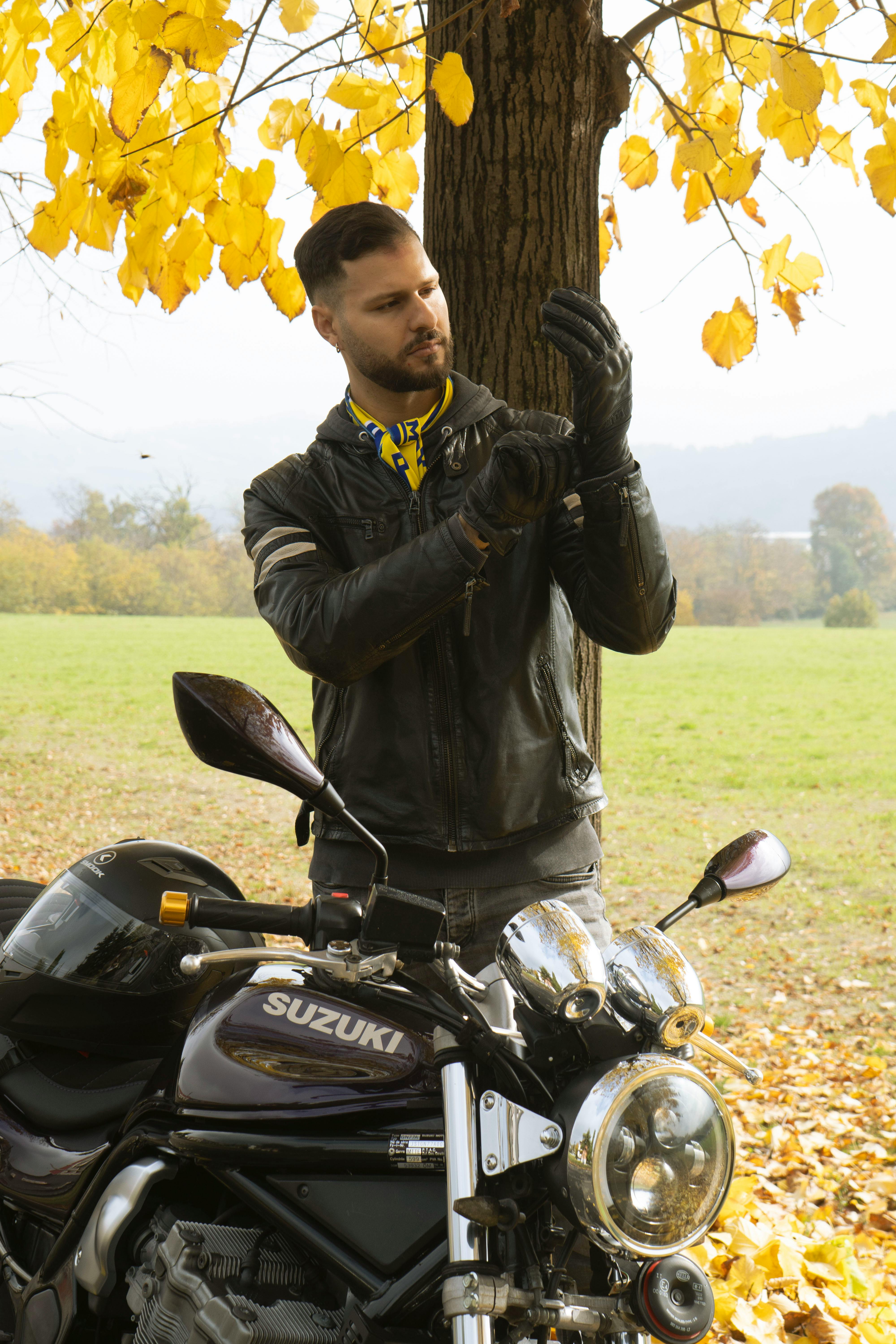 A male biker wearing gloves near a motorcycle under a tree during fall season.