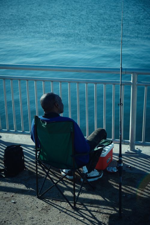 Free An adult man enjoys a calm day fishing by a lake with clear blue waters, embodying relaxation and nature's serenity. Stock Photo
