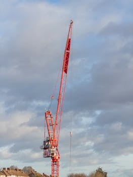 A towering red construction crane set against a cloudy sky in London.