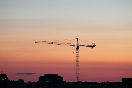 A construction crane silhouetted against a colorful sunset sky over a city skyline.