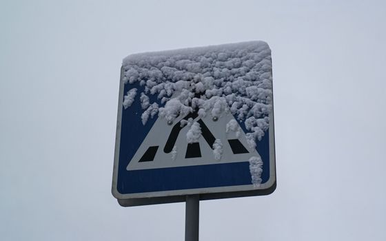 A pedestrian crossing sign partially covered with snow against a cloudy winter sky.