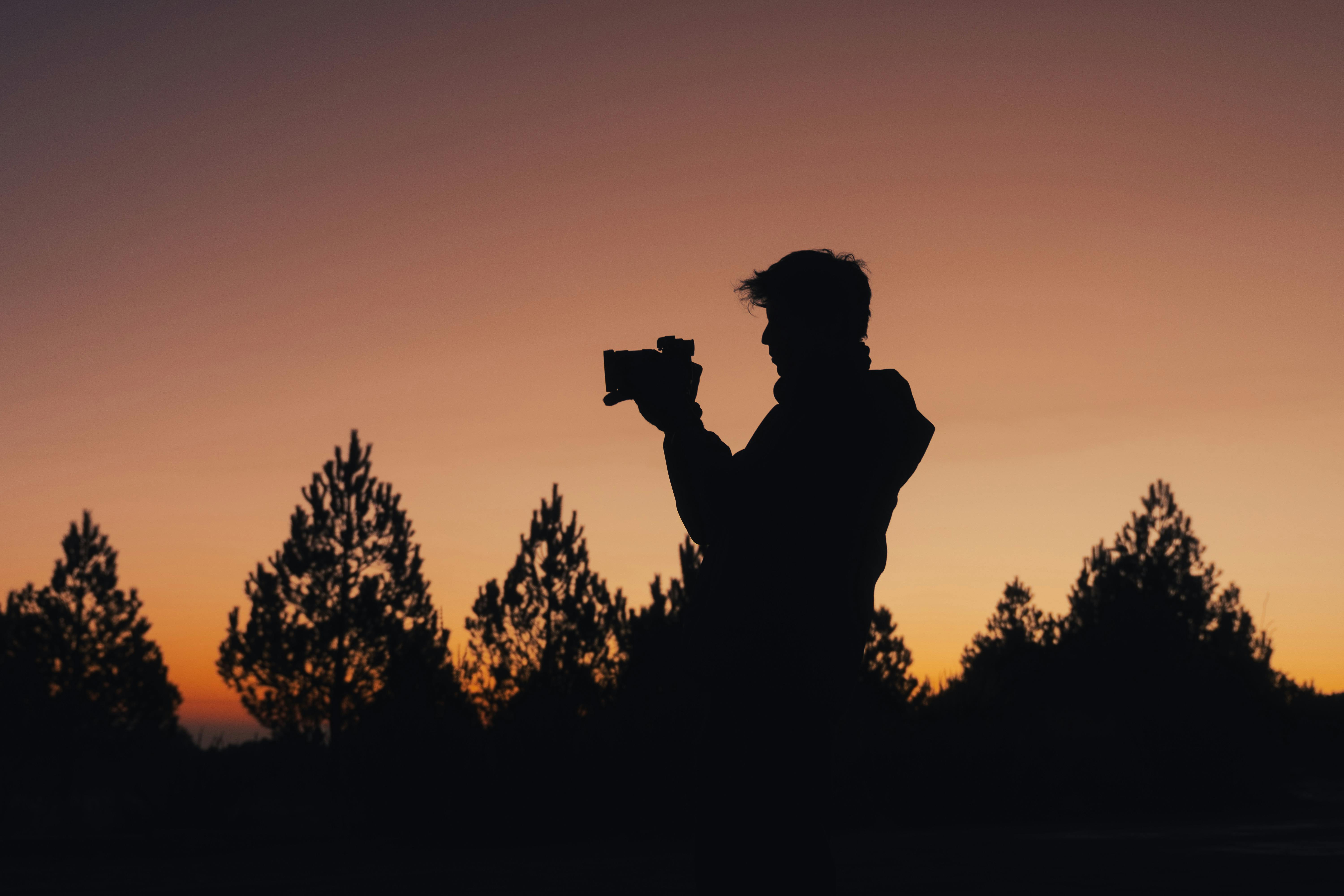 Gratis Silhuett av en fotograf som fanger solnedgangen i en skog med furutrær, og skaper et fredelig og dramatisk motiv. Arkivbilde