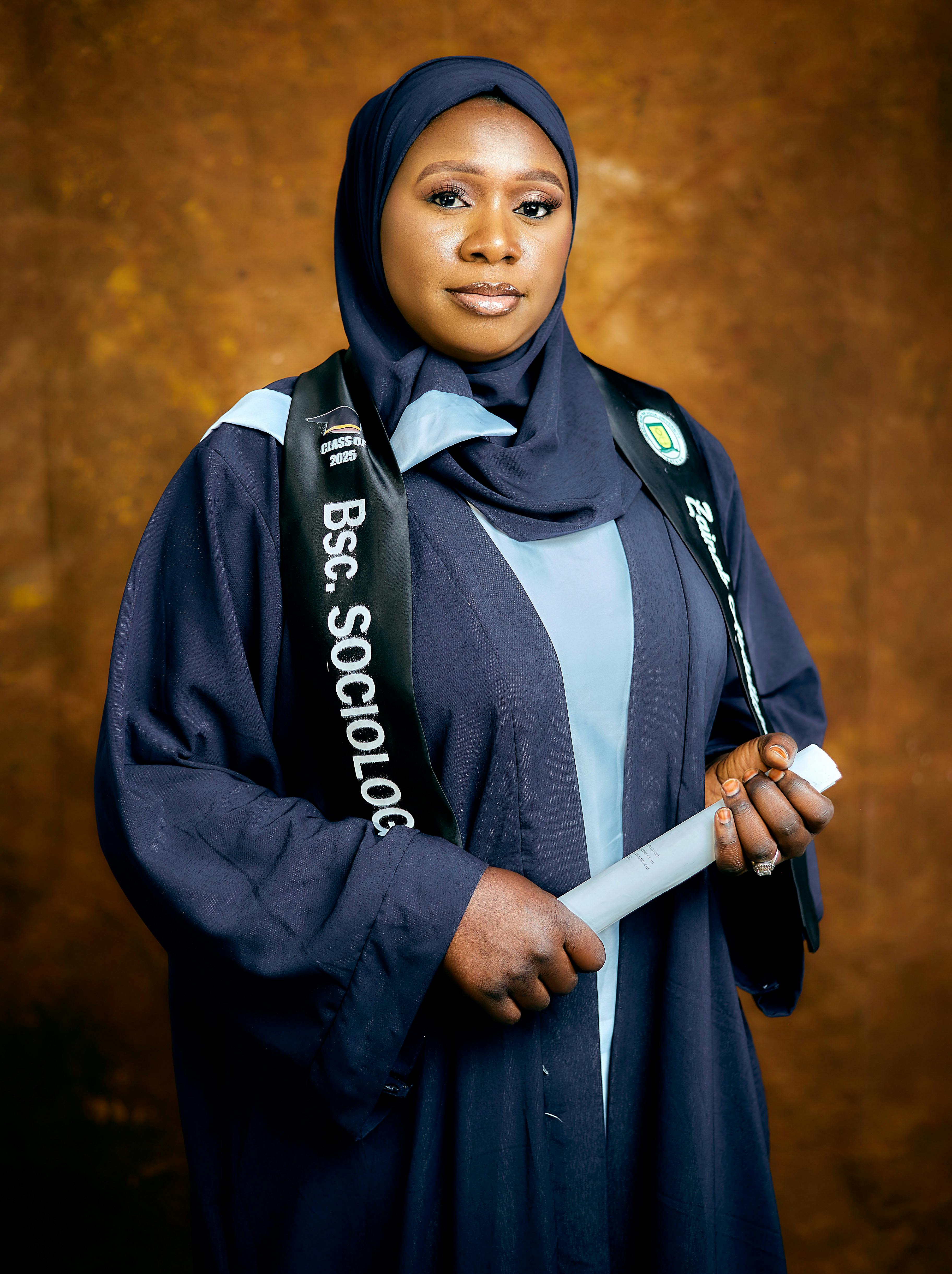 Confident female sociology graduate holding her diploma, wearing a hijab and graduation robe.