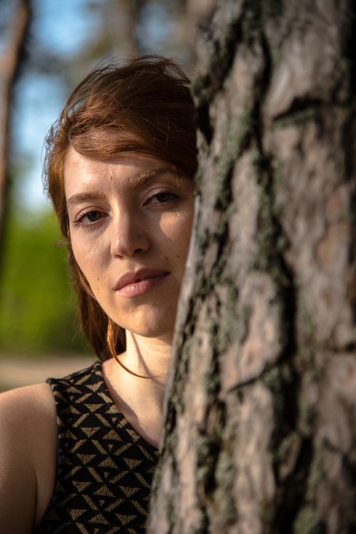 Free Close-up portrait of a woman with red hair leaning against a tree in a sunlit forest. Stock Photo