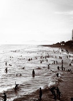 A busy beach with numerous people enjoying the ocean waves during sunset.