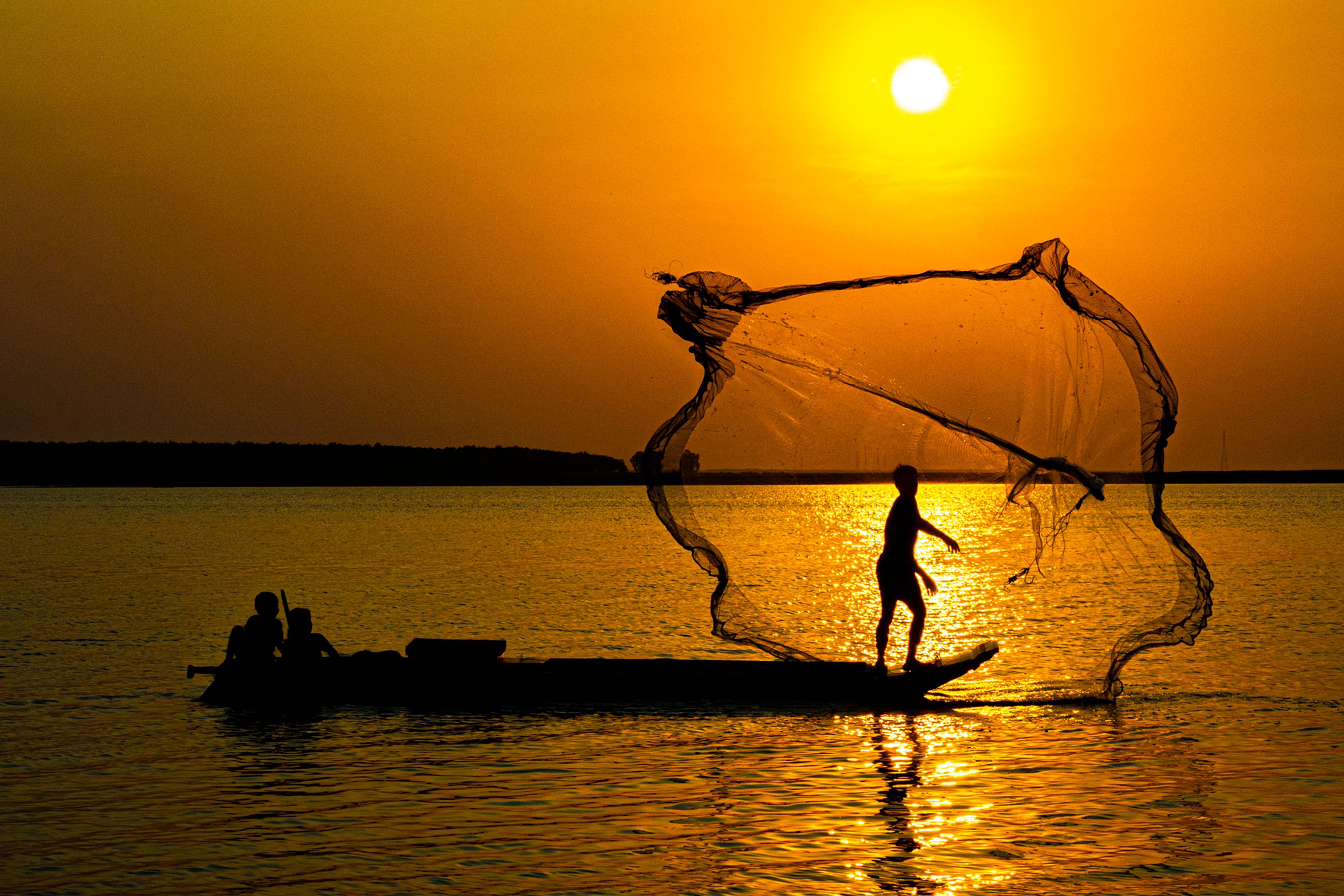 Silhouette of a fisherman casting a net on a calm lake at sunset, creating a serene and dramatic scene.