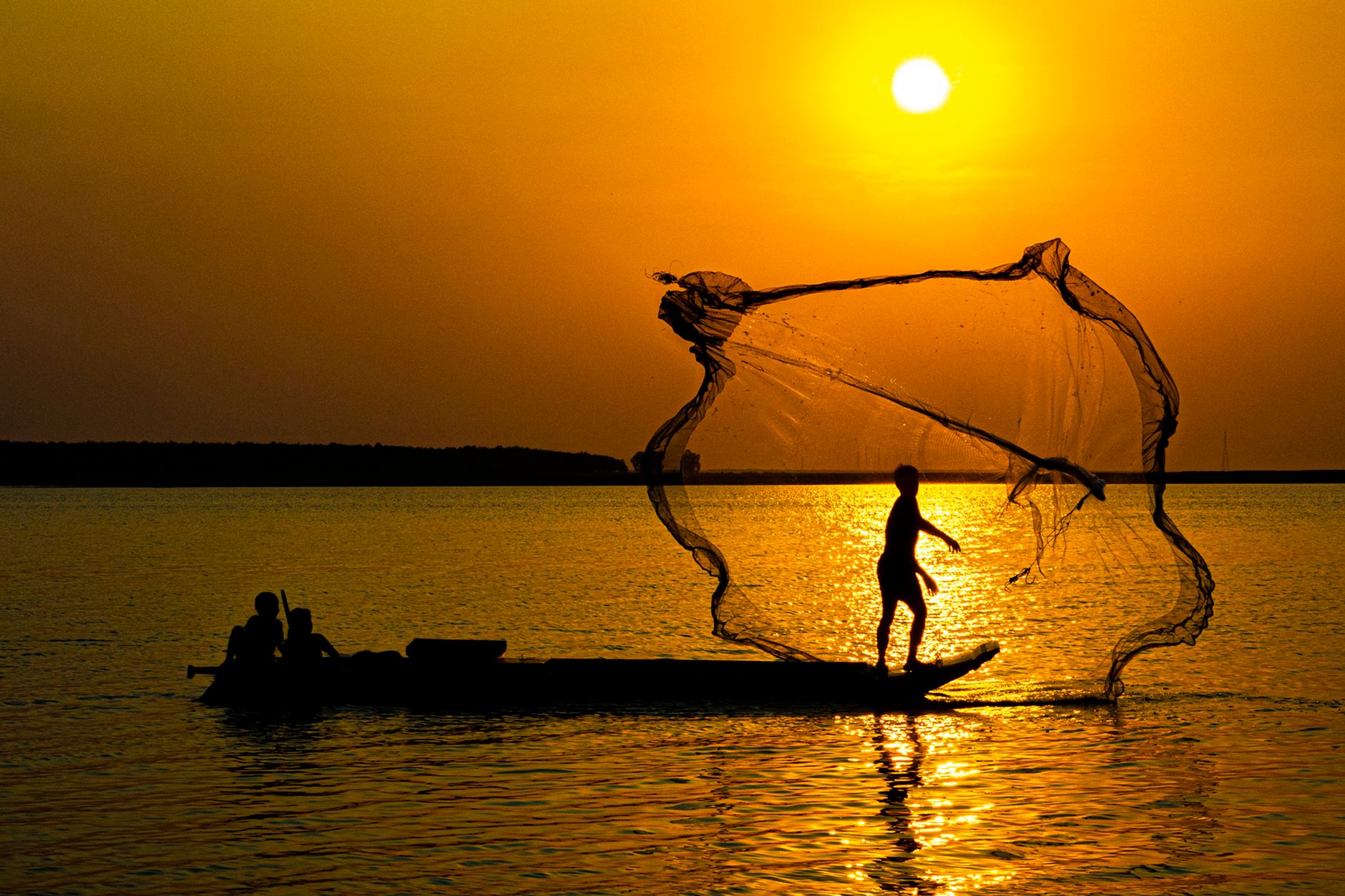 Silhouette of a fisherman casting a net on a calm lake at sunset, creating a serene and dramatic scene.