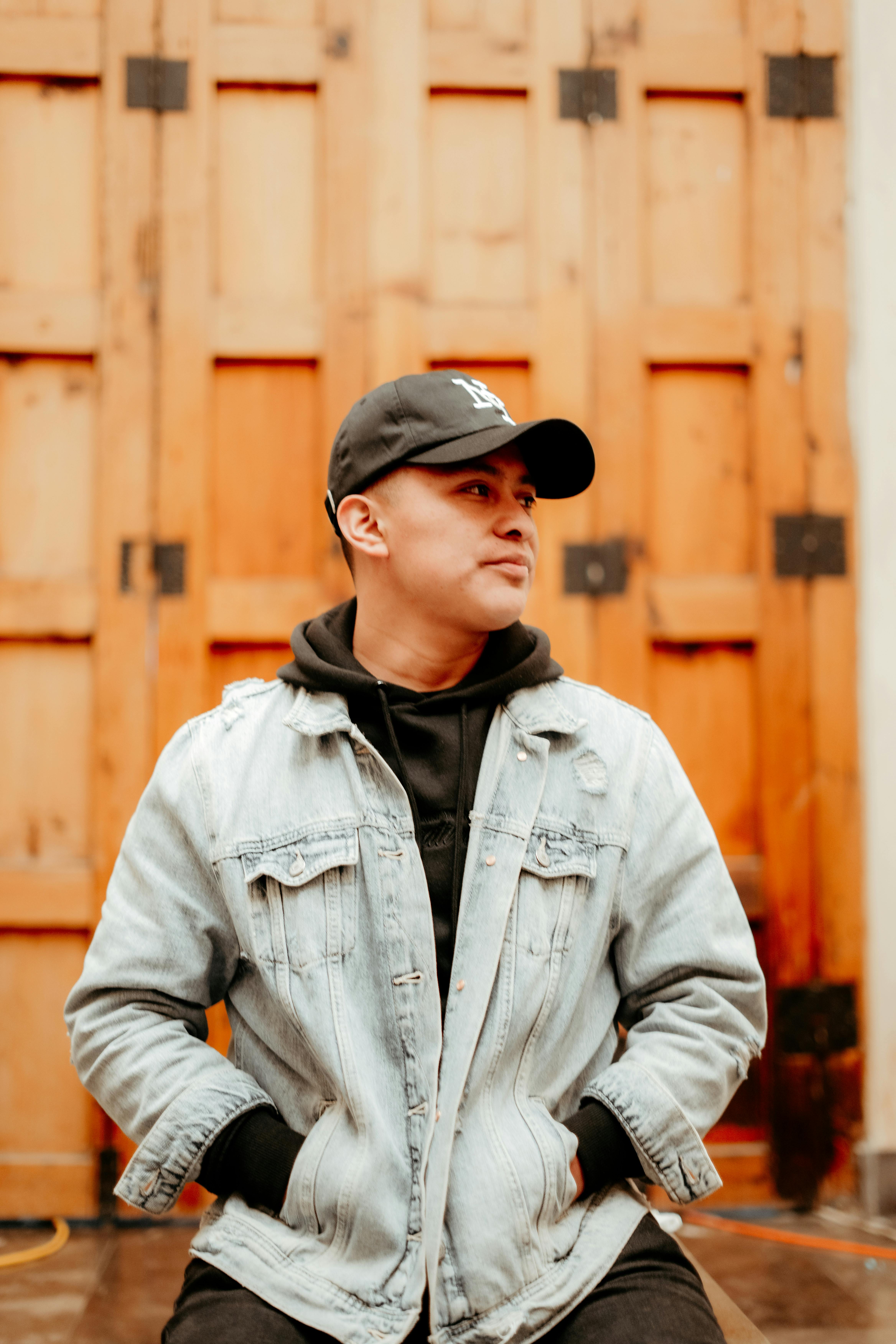 Man wearing denim jacket and cap sitting outdoors in front of wooden doors.