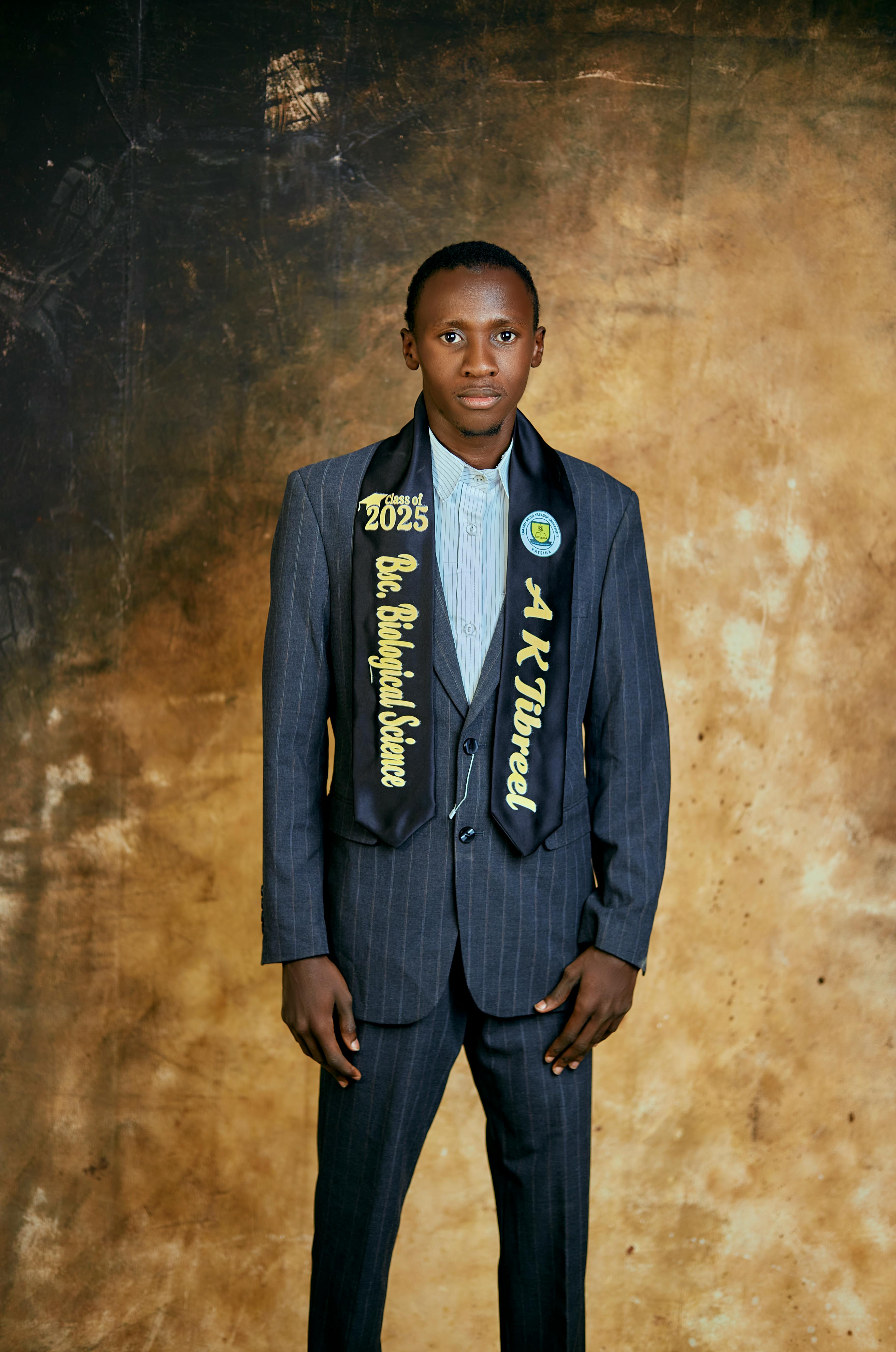 A young man in a suit poses for a graduation portrait with a sash in an indoor setting.