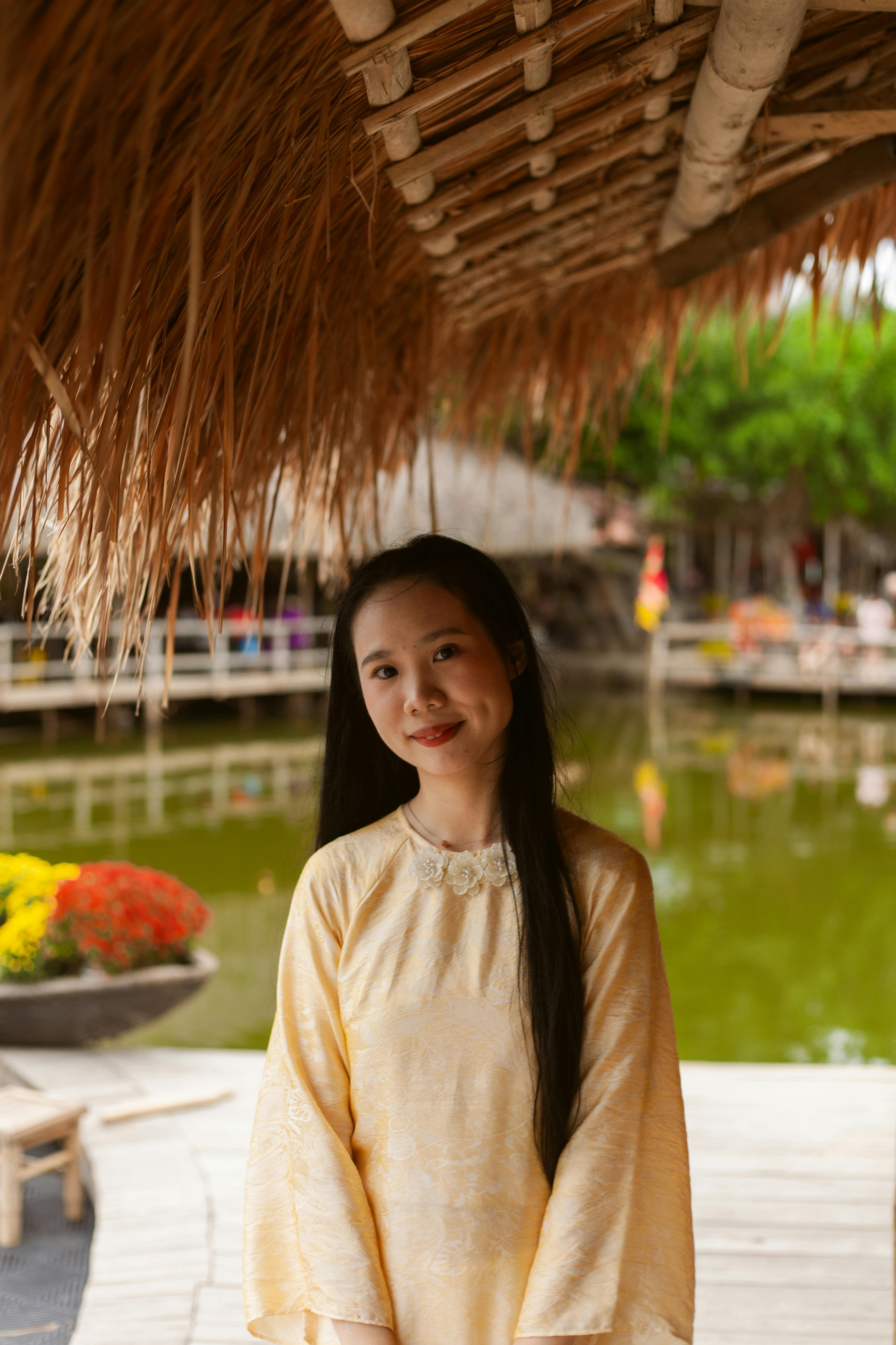 Gratis Una mujer joven con un vestido de seda vietnamita sonriendo bajo un techo de paja junto a un estanque. Foto de stock