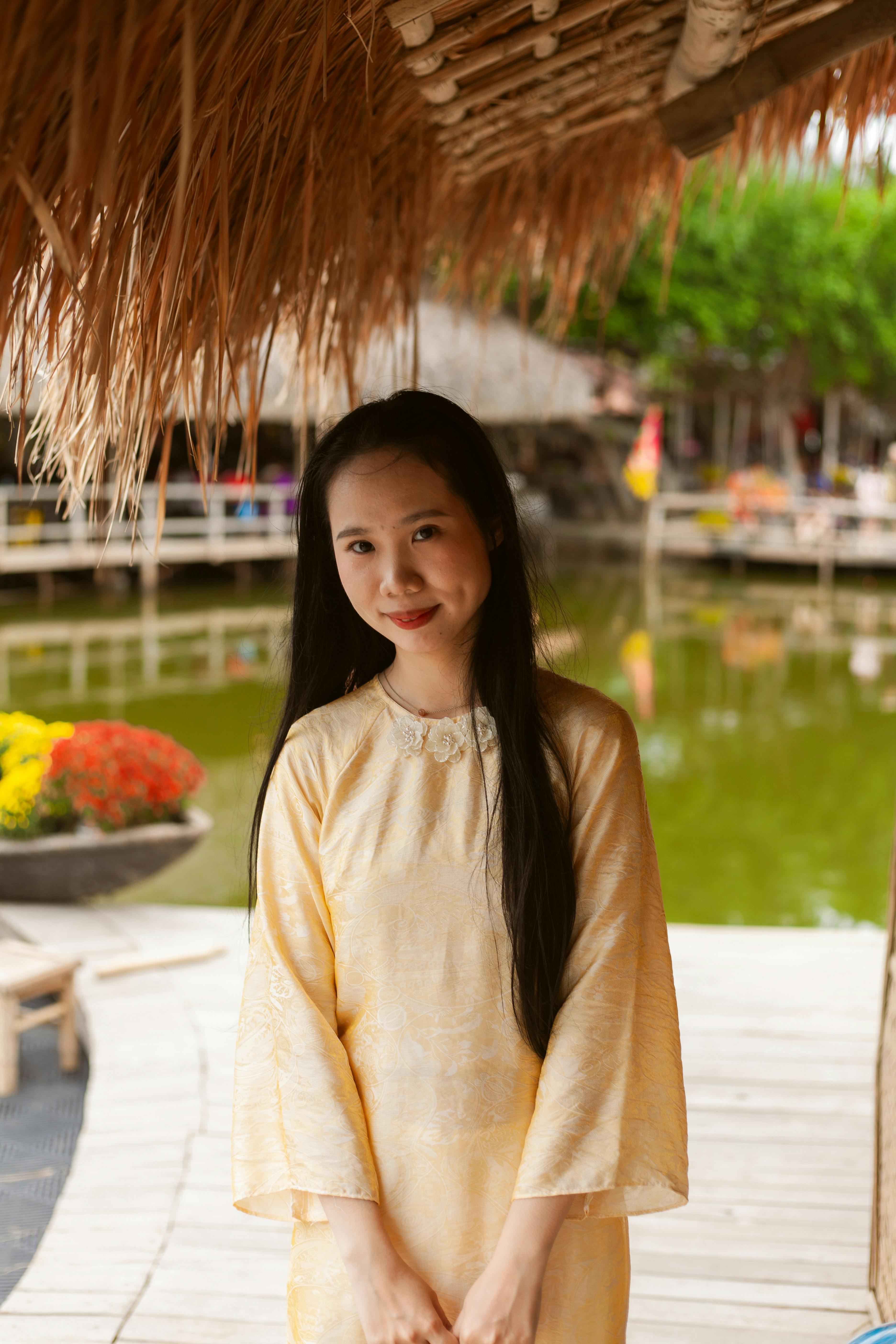 Gratis Mujer joven con vestido tradicional amarillo sonriendo bajo un techo de paja junto a un estanque. Foto de stock