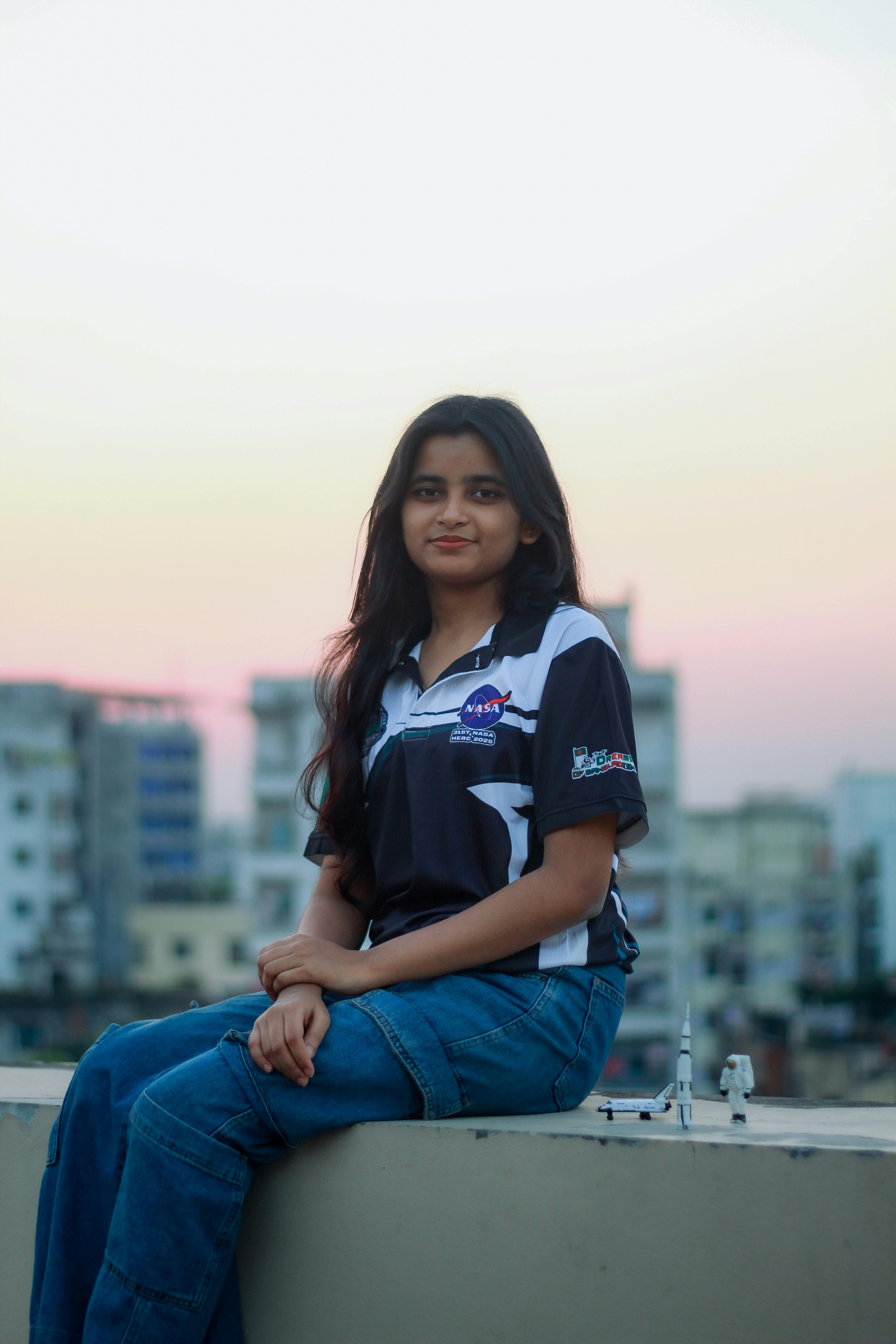 Gratis Una joven posa en una azotea de Dhaka al atardecer, vistiendo una camiseta con la temática de la NASA y con un paisaje urbano de fondo. Foto de stock