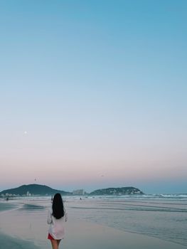 A woman stands on a serene beach at dusk with a pastel sky in the background.