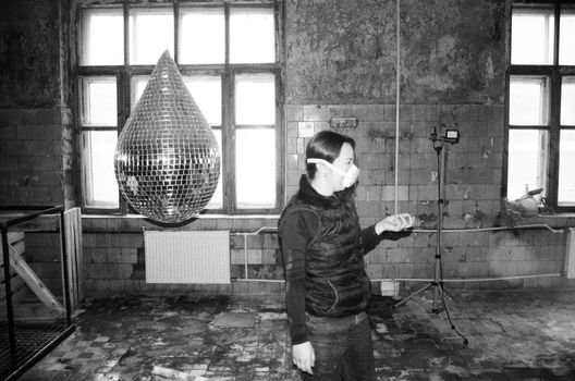 A woman in a mask stands indoors near a disco ball and exposed walls, creating a moody atmosphere.