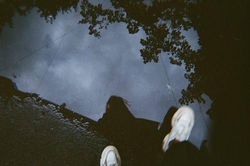 Free A person in sneakers walks past a puddle reflecting the sky and trees on a rainy day. Stock Photo