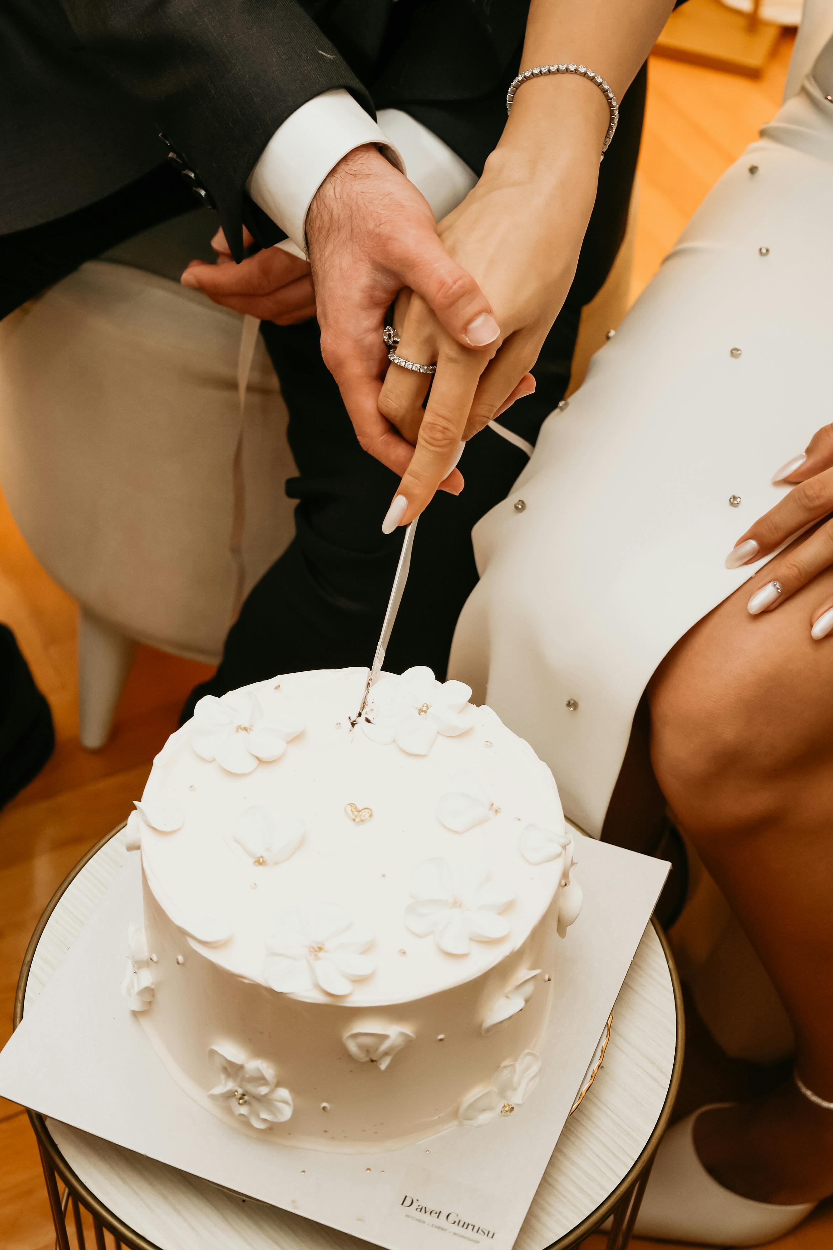 grátis Close-up das mãos de um casal cortando um bolo de casamento branco decorado com flores, simbolizando amor e celebração. Foto profissional