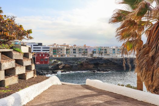 Scenic coastal view from a pathway in Las Palmas de Gran Canaria, Spain, featuring oceanside buildings and palm trees.