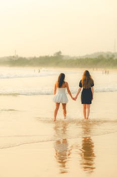 Two women walking hand in hand along a serene beach at sunset, reflecting friendship and tranquility.