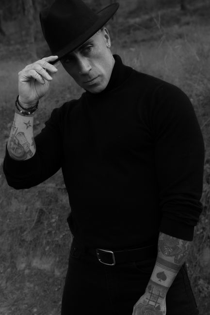 Black and white portrait of a tattooed man posing confidently in a black hat outdoors.