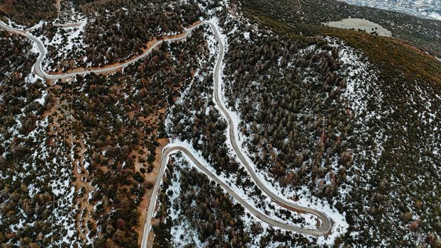 Winding mountain road cutting through a snowy forest landscape captured from above.