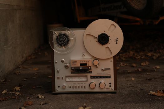 A classic AKAI reel-to-reel recorder on a leaf-strewn driveway, showcasing vintage audio technology.