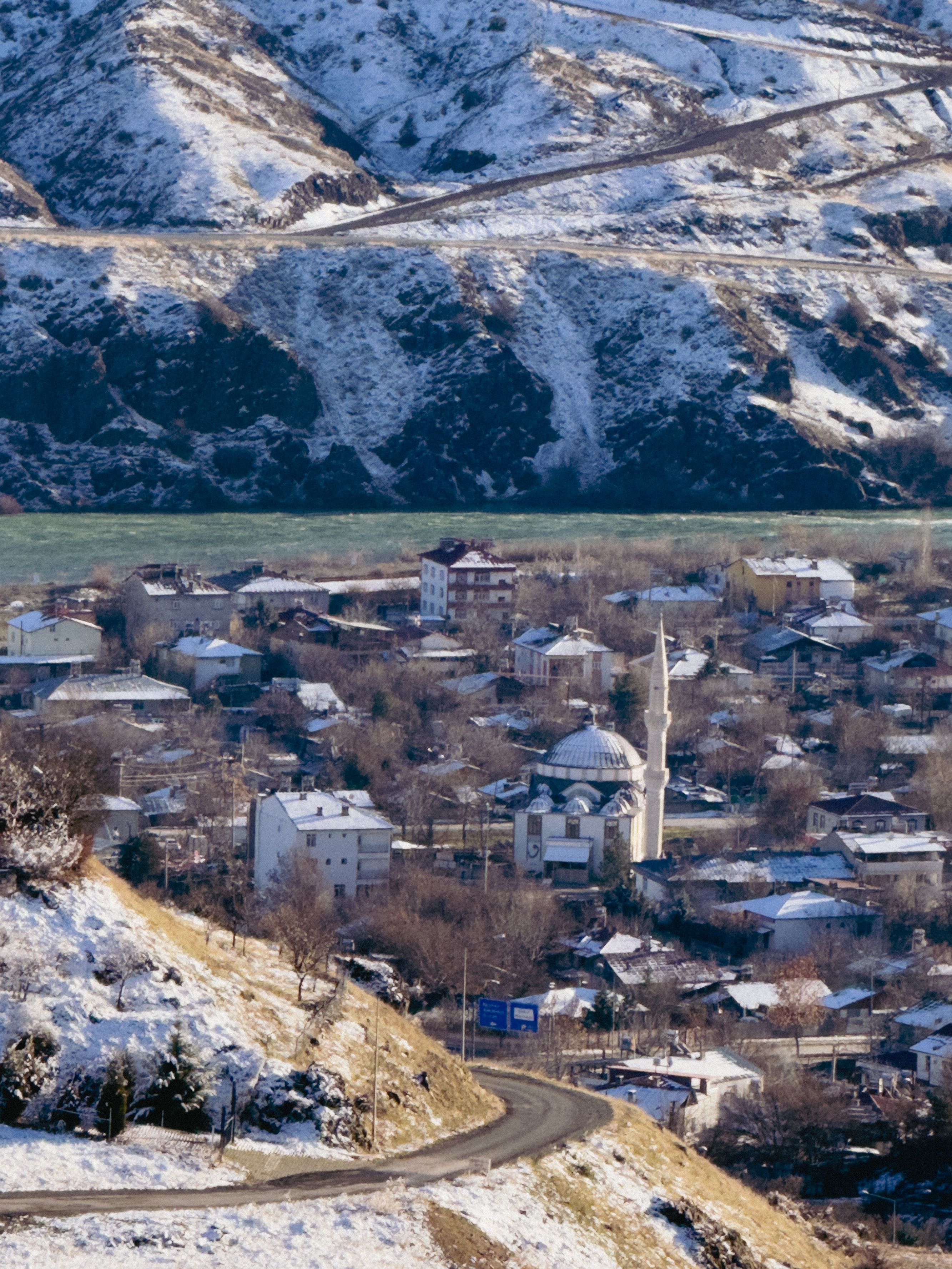 Village view of Palu with a mosque, snow-covered mountains, and a winding road in winter.