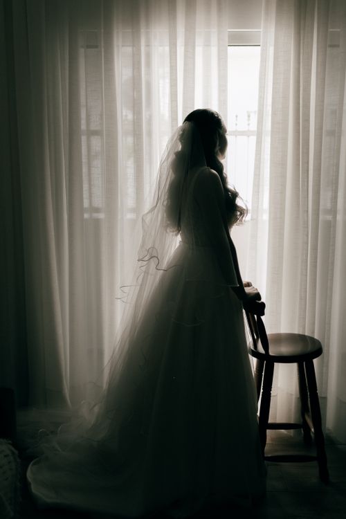 Free A beautiful bride in silhouette standing beside a window, creating a timeless and romantic scene. Stock Photo