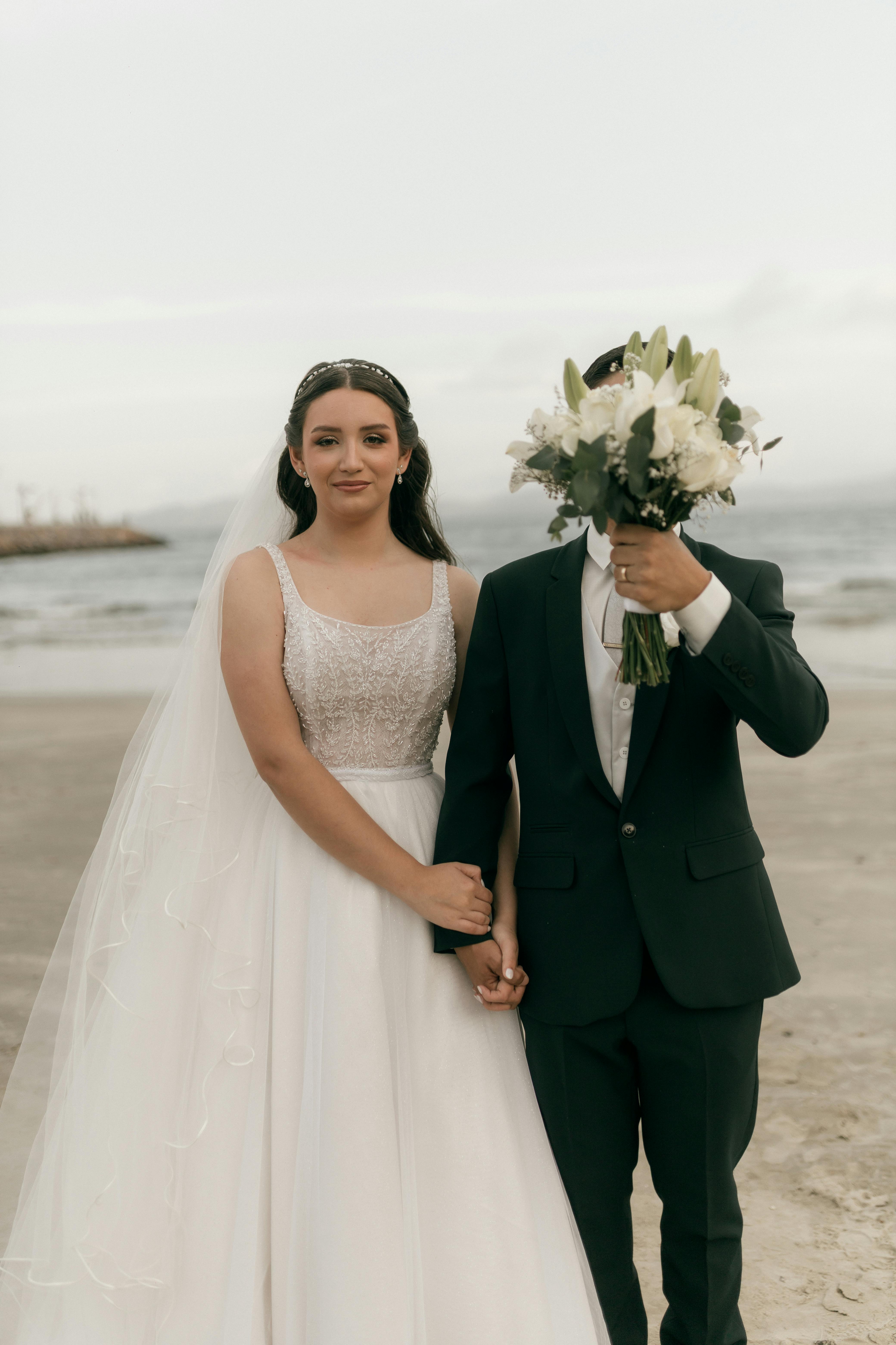 De franc Foto de casament a la platja amb parella, la núvia mostra elegància, el nuvi s'amaga amb humor darrere del ram. Foto d'estoc