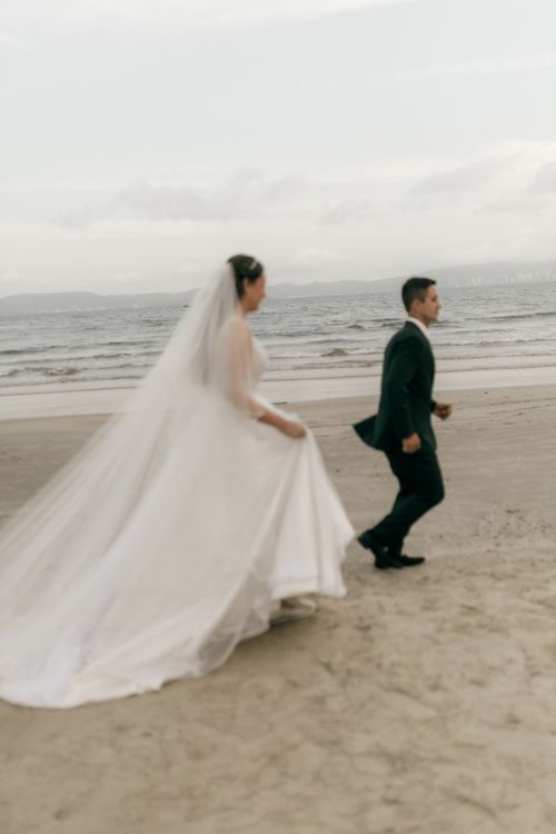 Free Bride and groom in wedding attire running on a beach with a serene ocean background. Stock Photo