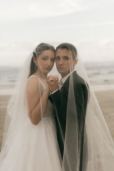 A couple in wedding attire poses romantically under a veil on a beach during the day.