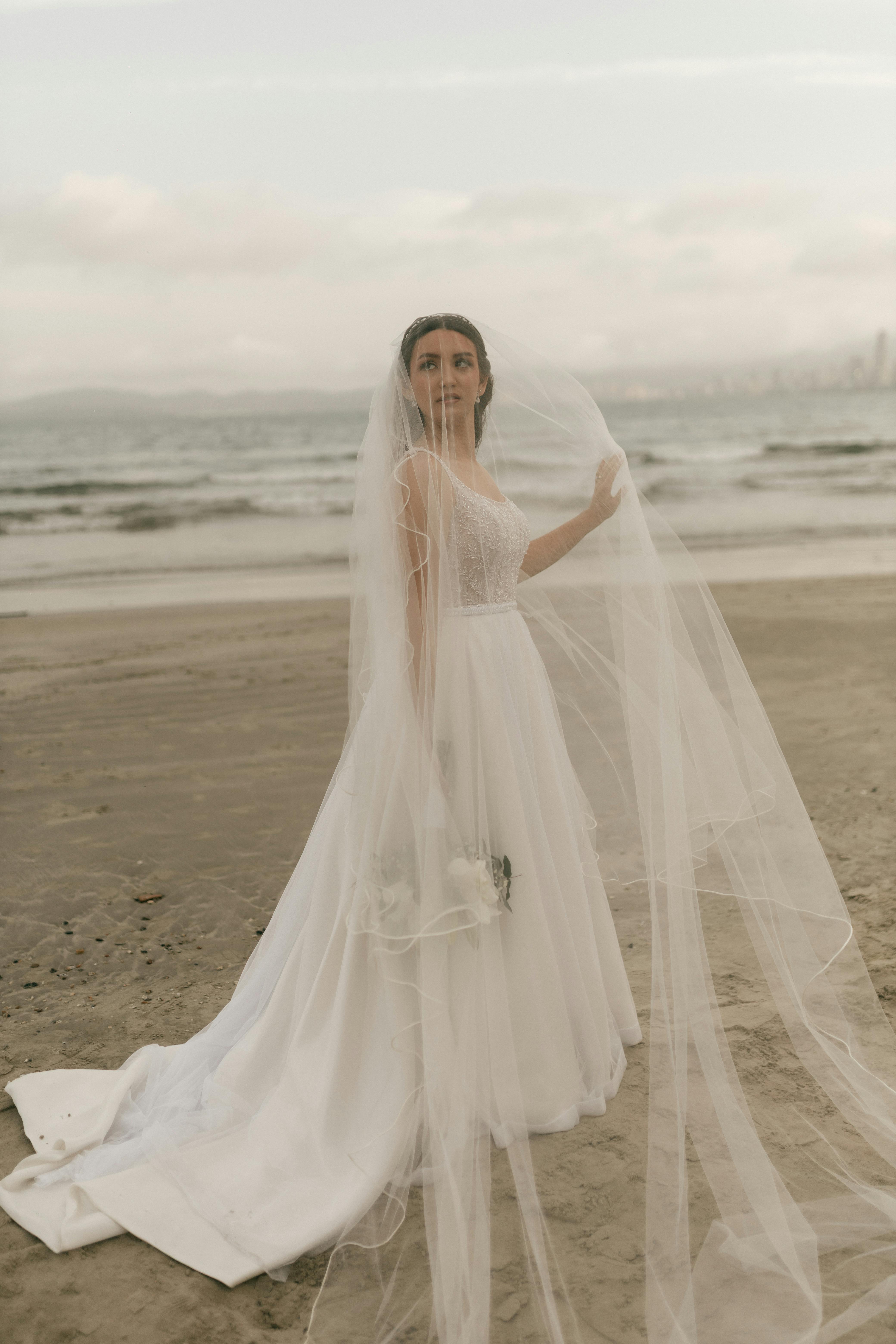 De franc La núvia amb un impressionant vestit blanc es troba elegant en una platja serena, amb el vel onejant amb la brisa. Foto d'estoc
