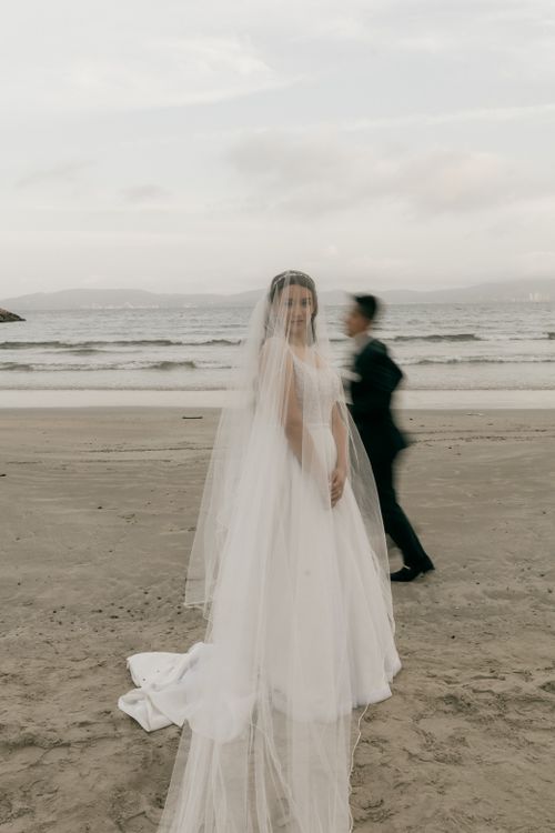 Free Bride in elegant wedding dress on a beach with groom in motion blur. Serene ocean backdrop. Stock Photo