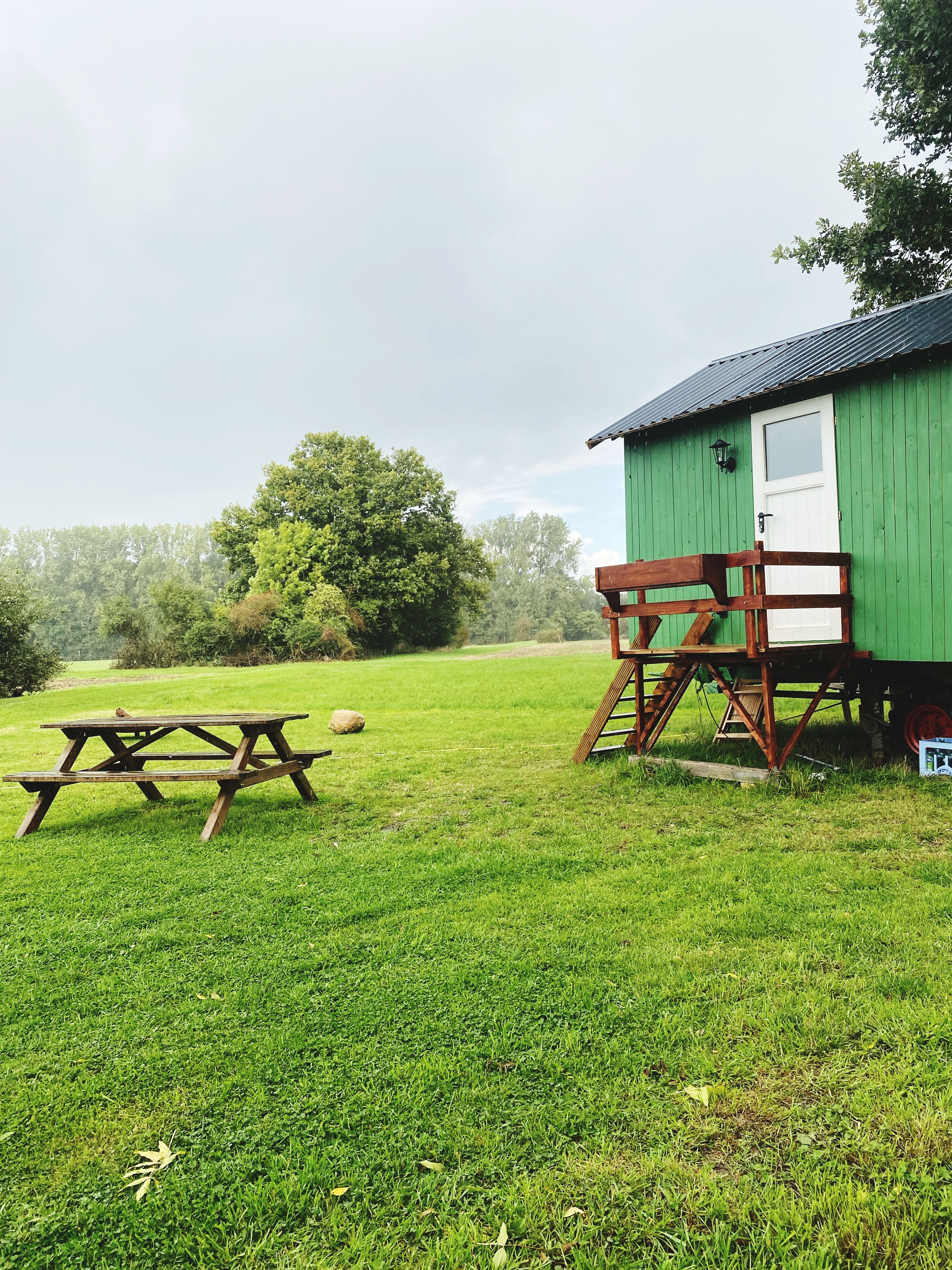 A green hut on a raised chassis with wooden steps, beside a picnic table on a grassy field