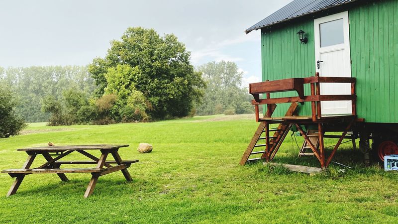 A green hut on a raised chassis with wooden steps, beside a picnic table on a grassy field