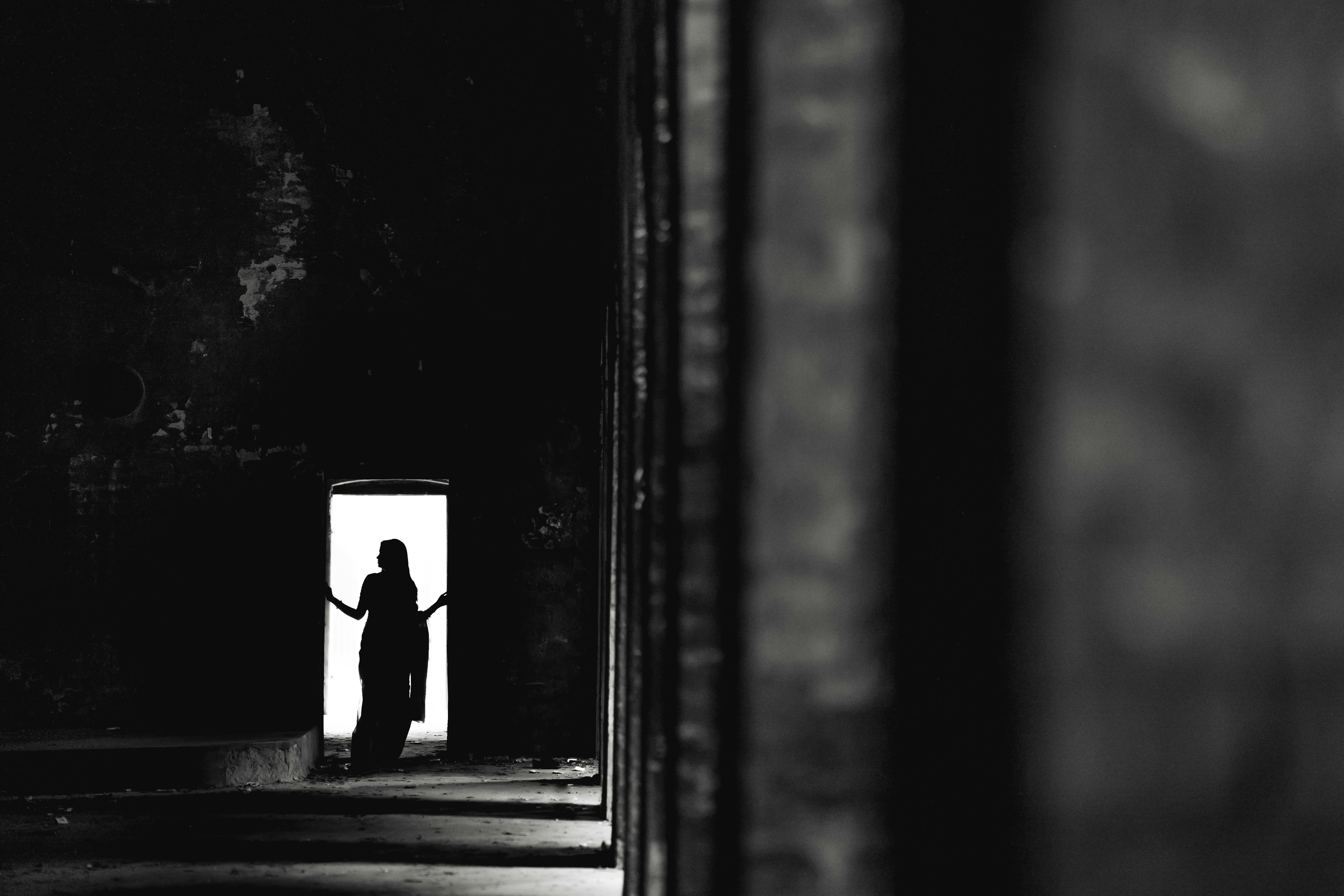 Gratis Una spettacolare silhouette in bianco e nero di una donna in piedi su una porta scarsamente illuminata, che evoca mistero e intrigo. Foto a disposizione