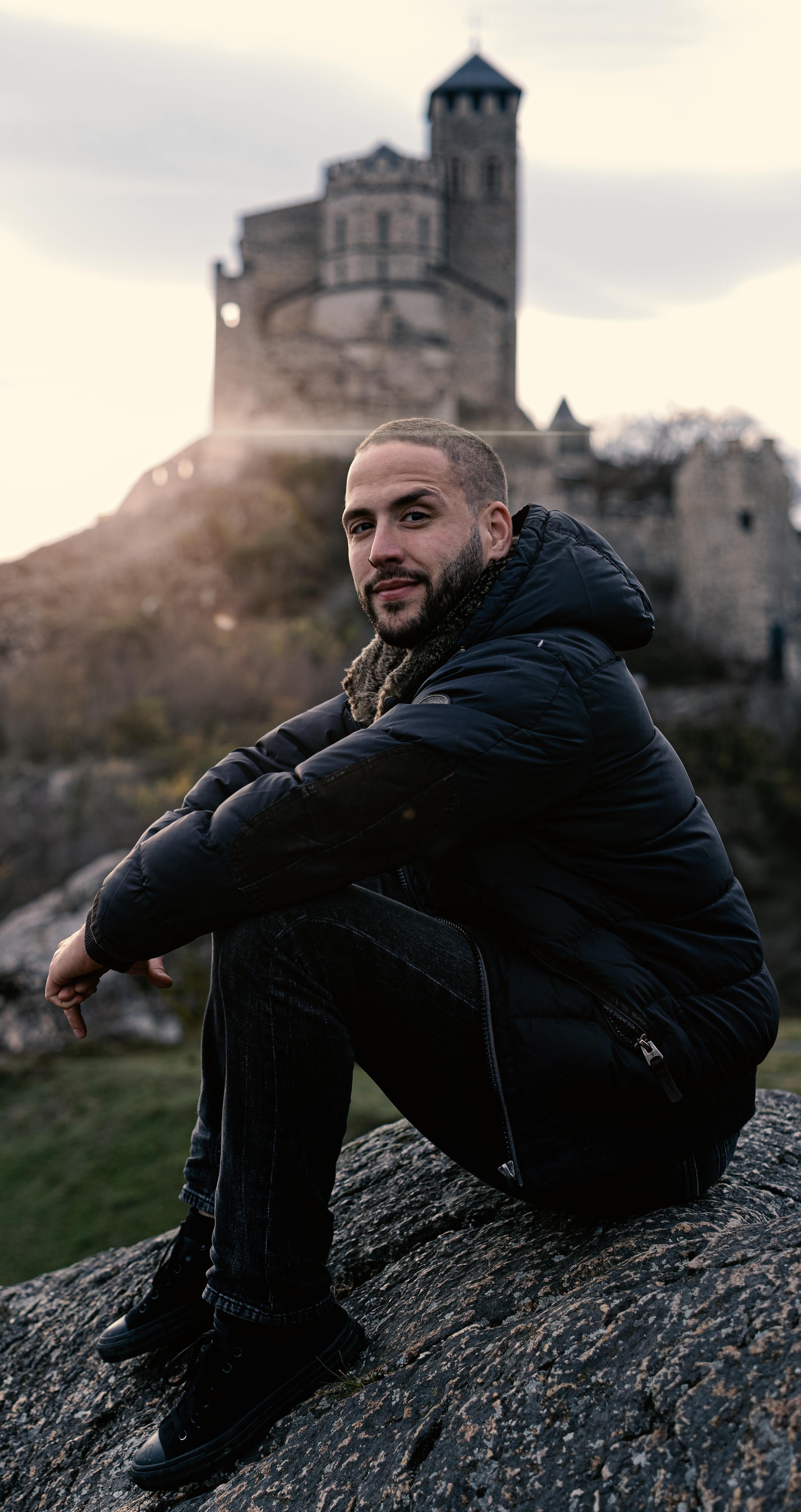 Man in winter clothing sits on a rock near a historic castle in Sion, Switzerland.