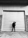 Grayscae Photo of Man Standing in Front of Closed Roll-up Door