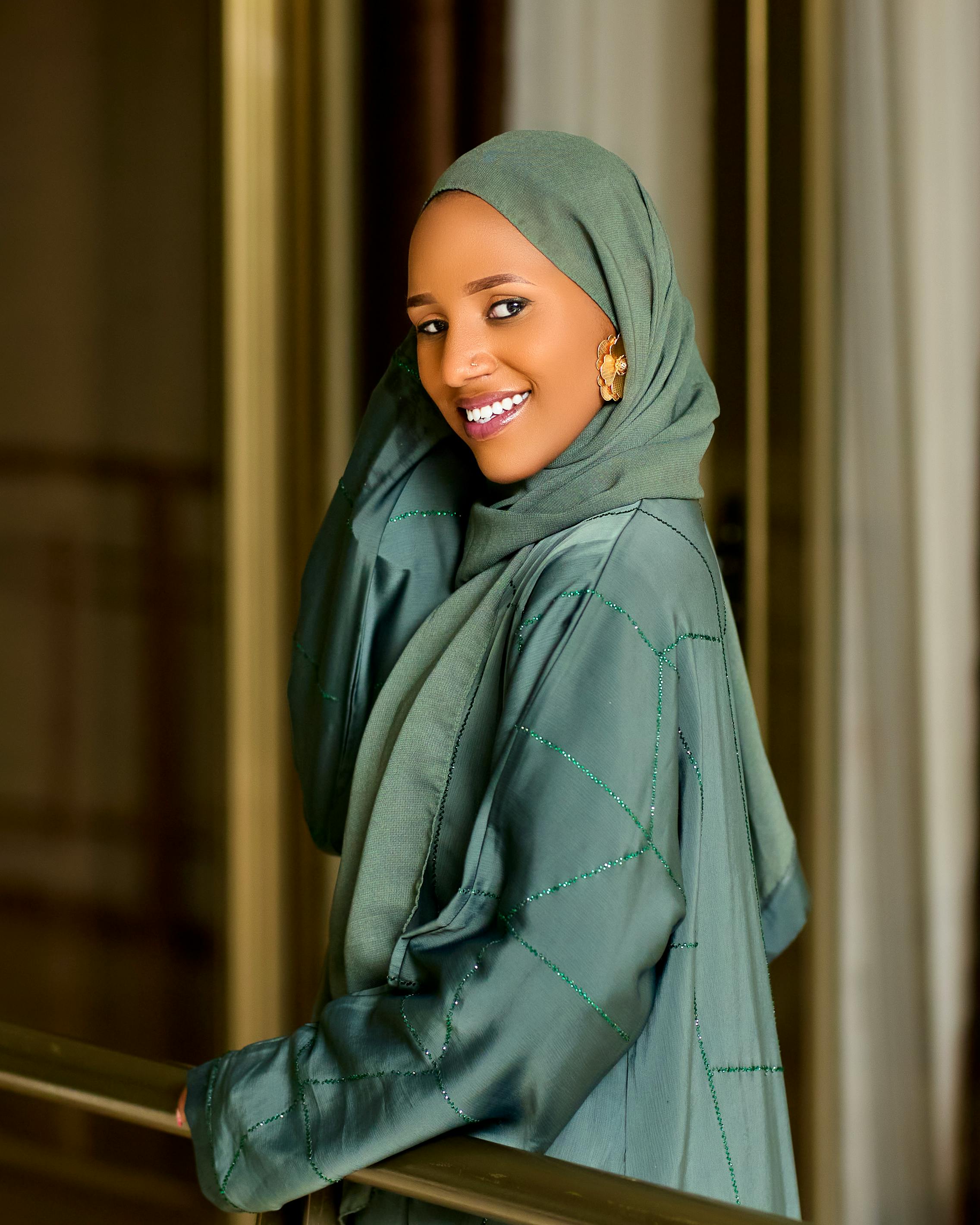 Portrait of a smiling woman in a green hijab and abaya indoors exuding elegance and warmth.
