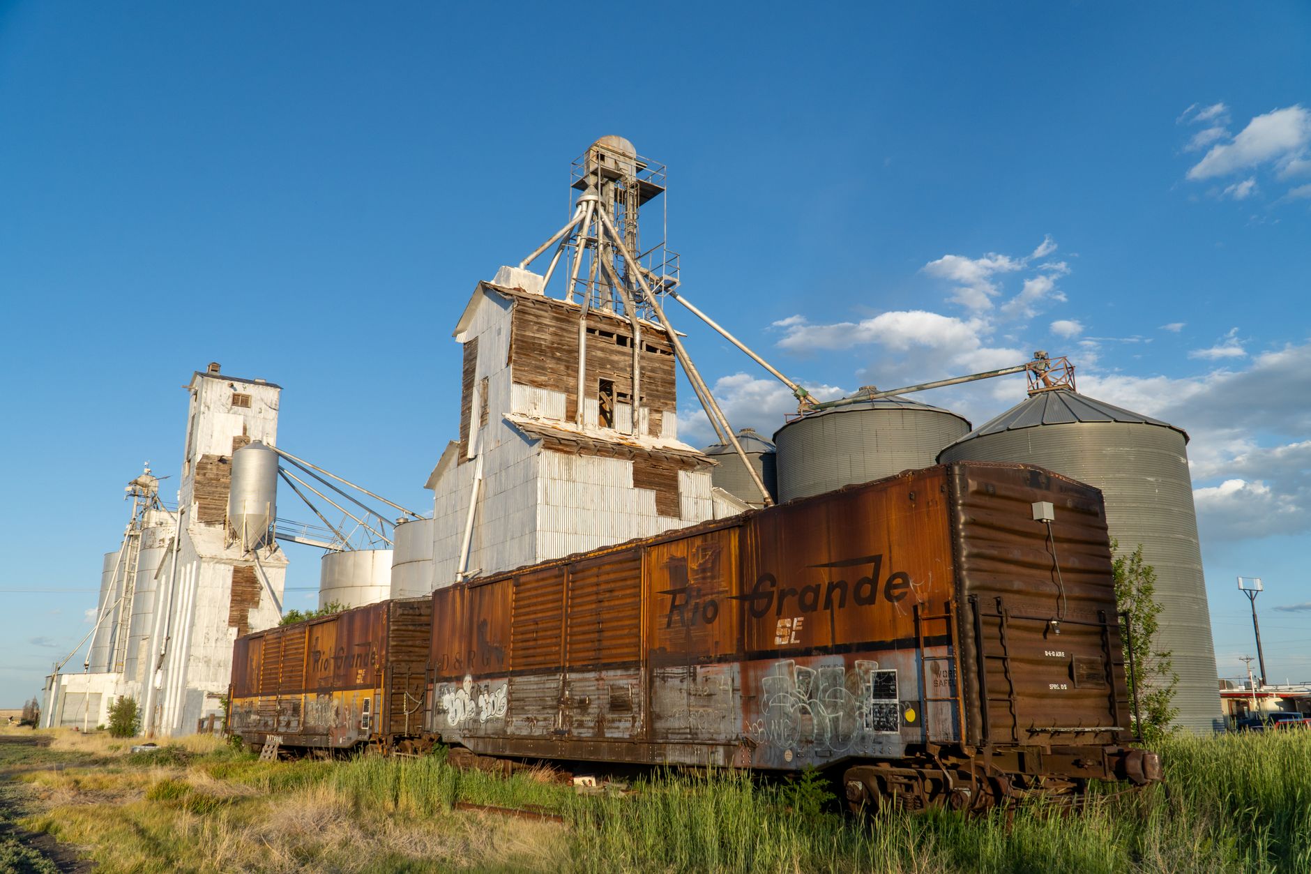 Rusty Rio Grande boxcars beside a grain elevator in Strasburg, Colorado under a clear blue sky.